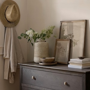Puede incluir: Una habitaci&oacute;n de tonos neutros con una c&oacute;moda gris oscuro, adornada con un sombrero de paja, estampados bot&aacute;nicos enmarcados y un jarr&oacute;n tejido lleno de vegetaci&oacute;n. Cuencos de madera y una pila de libros completan la decoraci&oacute;n.