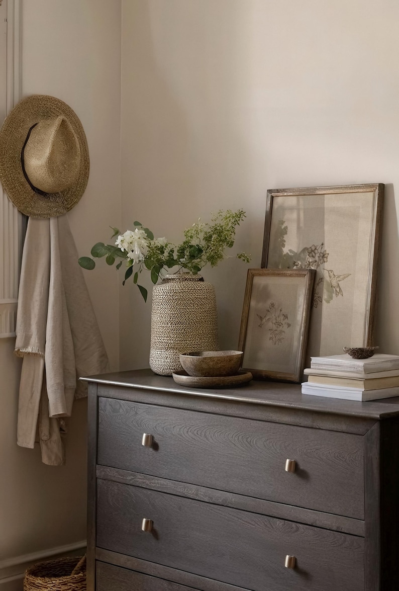 Puede incluir: Una habitaci&oacute;n de tonos neutros con una c&oacute;moda gris oscuro, adornada con un sombrero de paja, estampados bot&aacute;nicos enmarcados y un jarr&oacute;n tejido lleno de vegetaci&oacute;n. Cuencos de madera y una pila de libros completan la decoraci&oacute;n.