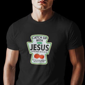Puede incluir: Camiseta negra con un gráfico en forma de botella de ketchup que dice "CATCH UP WITH JESUS" en verde y blanco. Debajo hay dos tomates rojos. La camiseta también tiene el texto "LETTUCE PRAKS AND RELLIN HIM" y "CO2 NE BLESSIS NEFSOM MY HEAD TO NO-7005".