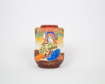 May include: A small, brown and white ceramic vase with gold accents. The vase is decorated with a colorful painting of a woman in traditional Japanese clothing.