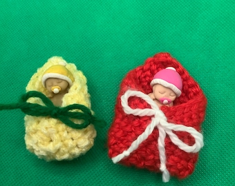 May include: Two crocheted baby doll sleeping bags, one yellow with a green bow and one red with a white bow. The dolls are wearing hats and have pacifiers in their mouths.