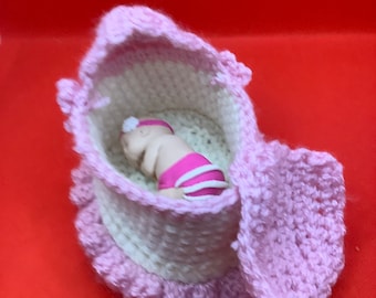 May include: A pink and white crocheted baby crib with a small doll inside. The crib has a ruffled edge and a removable lid.