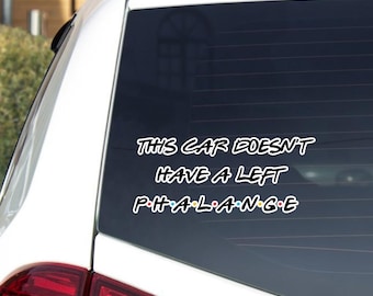 May include: A white car window with a black decal. The decal reads "THIS CAR DOESN'T HAVE A LEFT PHALANGE" in a white, stylized font. The text is arranged in three lines, with the word "PHALANGE" in a colorful font.