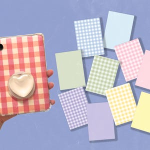 Pode incluir: Uma capa transparente para tablet com um padrão xadrez rosa e branco e um suporte em forma de coração. Há notas adesivas em tons pastel, incluindo azul, verde, rosa e amarelo. O fundo é azul claro.