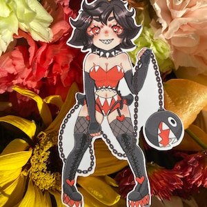 Może przedstawiać: Naklejka z postacią z kreskówki o czarnych włosach, w czerwono-czarnym stroju i koronie. Postać trzyma łańcuch z czarną kulą na końcu. Naklejka znajduje się na tle kwiatów.