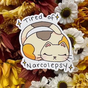 May include: A sticker featuring a cartoon cat curled up asleep. The cat is cream, brown, and orange. Text on the sticker reads "Tired of Narcolepsy" with star accents. The sticker is set against a background of yellow, white, and brown flowers.