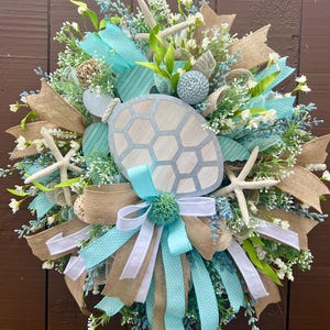May include: A decorative wreath featuring a central wooden turtle cutout with a geometric shell pattern, surrounded by burlap and light blue polka-dot ribbons, white starfish, small green foliage, and white floral accents on a circular frame.