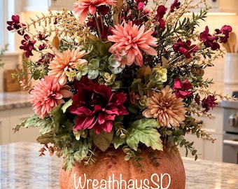 May include: A decorative arrangement of artificial flowers in shades of peach, deep red, and burgundy, featuring dahlias and greenery, set inside a textured orange pumpkin-shaped container with the text WreathhausSD printed on the front.
