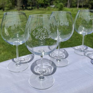 Puede incluir: Cuatro copas de vino transparentes con tallos largos están dispuestas sobre una superficie blanca. Cada copa está grabada con la palabra "Mom" y el nombre de la ciudad "Doylestown" o "Boylestown" en una fuente decorativa, con detalles florales.