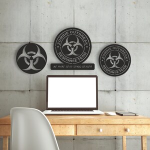May include: Three black metal wall decor signs with a biohazard symbol and the text "Zombie Outbreak Response Team" and "We make dead things deader". The signs are hanging on a gray concrete wall above a wooden desk with a laptop, a notebook, and a white mouse.