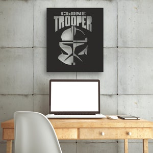 Puede incluir: Un letrero de metal blanco y negro con el texto "CLONE TROOPER" en letras blancas. El letrero presenta una imagen estilizada de un casco de soldado clon de Star Wars.