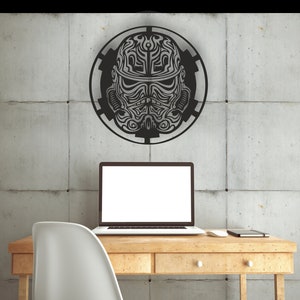 May include: A black metal wall decor featuring a stylized design of a Star Wars Stormtrooper helmet. The helmet is surrounded by a circular design with a gear-like pattern. The decor is mounted on a gray concrete wall above a wooden desk with a laptop and a chair.