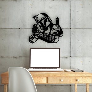 May include: A black metal wall decor featuring a Star Wars character, Boba Fett, holding a blaster rifle. The decor is mounted on a gray concrete wall above a wooden desk with a laptop, a chair, and a notebook.