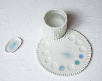 May include: White ceramic palette with 12 wells for mixing paint. The palette has a raised edge and a textured surface. A white ceramic cup with a textured surface sits on top of the palette.