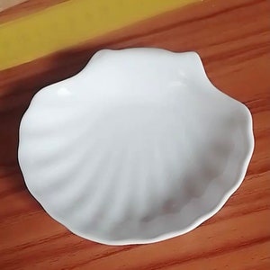 Könnte beinhalten: Eine weiße, muschelförmige Schale aus Keramik. Die Schale hat einen geriffelten Rand und eine glatte, glänzende Oberfläche.