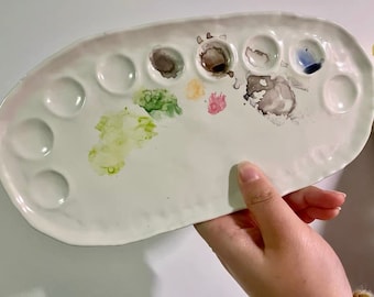 May include: A white ceramic paint palette with 10 circular wells. The palette has been used and has various colors of paint in the wells, including green, yellow, pink, brown, and blue.