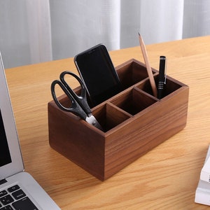May include: A wooden desk organiser with four compartments. The organiser is brown and has a phone, scissors, a pen, and a pencil inside.