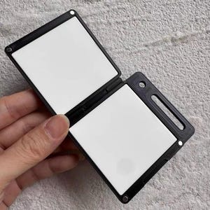 Op de afbeelding: Een zwart, scharnierend, vierkant object met twee witte panelen. Het object wordt opengehouden, waardoor de witte oppervlakken zichtbaar worden. Het frame heeft een rechthoekige uitsparing en een klein rond gat. Het object lijkt van plastic te zijn.