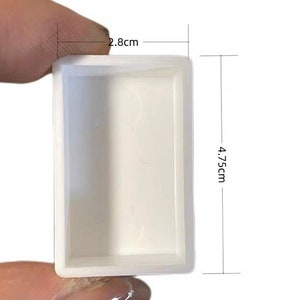 Może przedstawiać: Mały, prostokątny biały pojemnik o szerokości 2,8 cm i wysokości 4,75 cm trzymany jest między palcami. Pojemnik ma gładkie, matowe wykończenie i prosty, minimalistyczny design. Wymiary są wyraźnie zaznaczone.