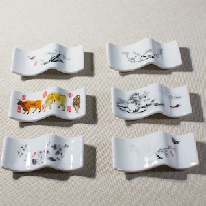 Könnte beinhalten: Set aus sechs weißen Keramik-Essstäbchenhaltern. Jeder Halter zeigt ein einzigartiges asiatisch inspiriertes Design, darunter Landschaftsbilder und Tierillustrationen. Die Halter haben eine wellenförmige Form und sind ideal zum Halten von Essstäbchen.