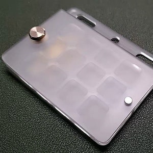 May include: A clear plastic card holder with a grid of nine compartments. The holder has a silver metal button on the side and a gold metal button on the top.