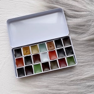 Puede incluir: Un estuche de acuarelas de metal blanco abierto, con 20 pastillas de pintura cuadradas. Los colores incluyen tonos de negro, marrón, verde y rojo. El conjunto se muestra sobre una superficie texturizada blanca y gris.