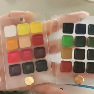 May include: Two clear plastic palettes, each containing 12 watercolour paint pans. The palette on the left features a range of warm colours, including white, yellow, orange, red, and brown. The palette on the right features a range of cool colours, including black, green, blue, and brown.