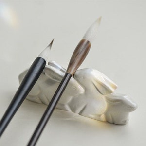 以下が含まれることがあります： 白いセラミック製のウサギの形をしたホルダーに、黒と茶色の2本の書道筆が置かれています