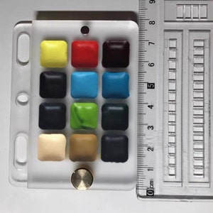 May include: A white plastic palette with 15 small squares filled with different colours of watercolour paint. The palette has a brass knob on the bottom right corner.