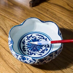 Puede incluir: Un tintero de cerámica blanco y azul con borde festoneado. El plato presenta un diseño floral azul en el centro y a lo largo del borde. Un pincel de caligrafía rojo y blanco descansa dentro del plato, listo para usar.