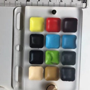 May include: A white plastic palette with 12 watercolour paint pans. The pans are arranged in four rows of three. The colours are yellow, red, black, blue, green, brown, beige, and grey.