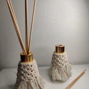 May include: Two white diffuser bottles with gold lids and gold trim. The bottles are wrapped in macrame and have tassels. Wooden diffuser sticks are in the bottles and on the table.
