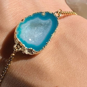 May include: A gold chain bracelet with a turquoise agate geode pendant. The geode has a white interior and a gold rim.