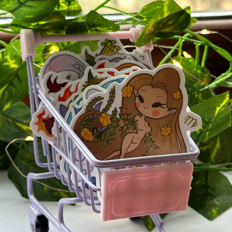 K&ouml;nnte beinhalten: Ein Miniatur-Einkaufswagen gef&uuml;llt mit bunten Stickern mit astrologischen Zeichen. Die Sticker sind mit Comicfiguren und floralen Designs illustriert. Ein Sticker zeigt eine Comic-Jungfrau mit langen braunen Haaren und einer Blumenkrone.