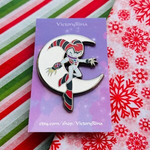 May include: A silver-colored pin featuring a cartoon character standing on a crescent moon. The character is white with red, black, and purple accents. The pin is on a purple card with the text "VictoryRina" and "etsy.com/shop/VictoryRina".
