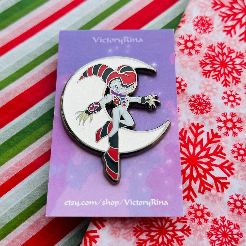 May include: A silver-colored pin featuring a cartoon character standing on a crescent moon. The character is white with red, black, and purple accents. The pin is on a purple card with the text "VictoryRina" and "etsy.com/shop/VictoryRina".
