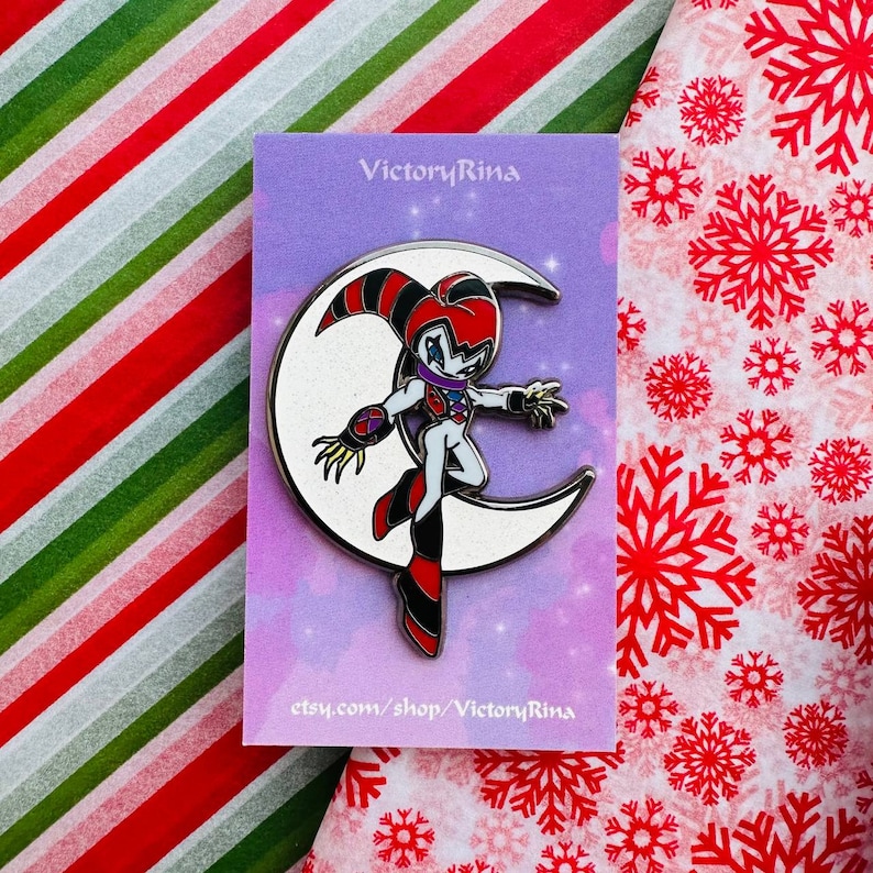 May include: A metal pin featuring a cartoon character standing on a crescent moon. The character has a red, black, white, and purple color scheme. The pin is on a purple card with the text "VictoryRina" and "etsy.com/shop/VictoryRina".