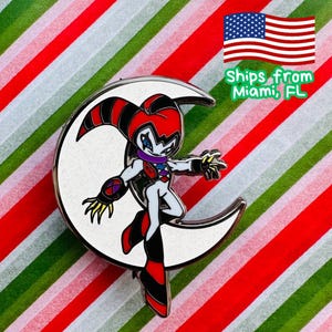 May include: Enamel pin featuring a character in a jester-like outfit with red, black, and white details, posed on a crescent moon. The pin has a silver border and is set against a striped background of red, white, and green.