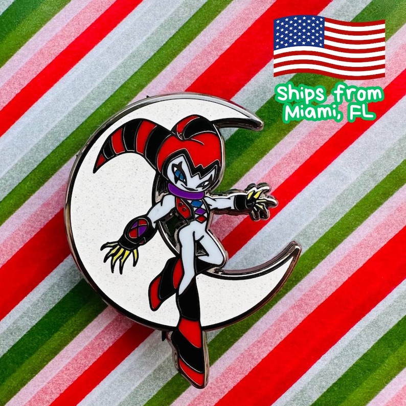 May include: Enamel pin featuring a character in a jester-like outfit with red, black, and white details, posed on a crescent moon. The pin has a silver border and is set against a striped background of red, white, and green.
