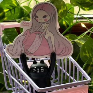 K&ouml;nnte beinhalten: Eine schwarze Katzenfigur mit wei&szlig;en Augen befindet sich in einem lila Einkaufswagen. Ein Aufkleber einer pinkhaarigen Frau, die einen rosa Wasserkrug mit dem Sternzeichen Wassermann h&auml;lt, befindet sich oben auf dem Wagen.