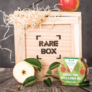 May include: A wooden crate with the words "RARE BOX" printed on the front. The crate is filled with shredded paper and a red apple. A small jar of smoked pork, apple & cider p&acirc;t&eacute; is sitting on a wooden table in front of the crate.