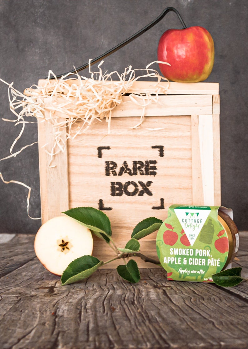 May include: A wooden crate with the words "RARE BOX" printed on the front. The crate is filled with shredded paper and a red apple. A small jar of smoked pork, apple & cider p&acirc;t&eacute; is sitting on a wooden table in front of the crate.