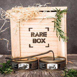 May include: Two cans of canned meat in a wooden crate with the words "RARE BOX" printed on the front. The cans are labeled "Hirschkalbfleisch Canned in its own juice" and feature images of a moose and a deer.