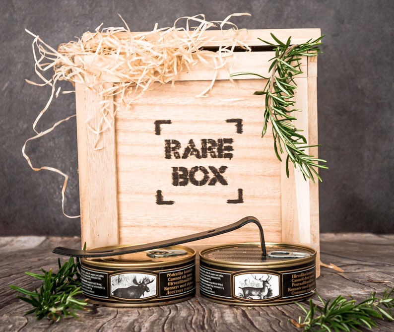 May include: Two cans of canned meat in a wooden crate with the words "RARE BOX" printed on the front. The cans are labeled "Hirschkalbfleisch Canned in its own juice" and feature images of a moose and a deer.