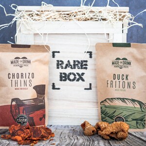 May include: A white wooden crate with the words "RARE BOX" printed on the front. The crate contains two bags of snacks, one with chorizo thins and the other with duck fritons. Both bags have the words "MADE FOR DRINK" printed on them.