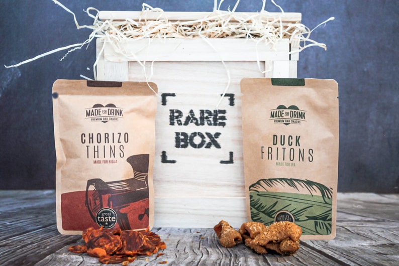 May include: A white wooden crate with the words "RARE BOX" printed on the front. The crate contains two bags of snacks, one with chorizo thins and the other with duck fritons. Both bags have the words "MADE FOR DRINK" printed on them.