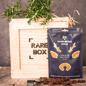 May include: A wooden crate with the words "Rare Box" printed on the front. The crate contains a bag of venison biltong from the brand "Wholesome Wolf". The bag is blue with a deer head logo and the text "Venison Biltong Classic". The biltong is also visible outside of the bag.