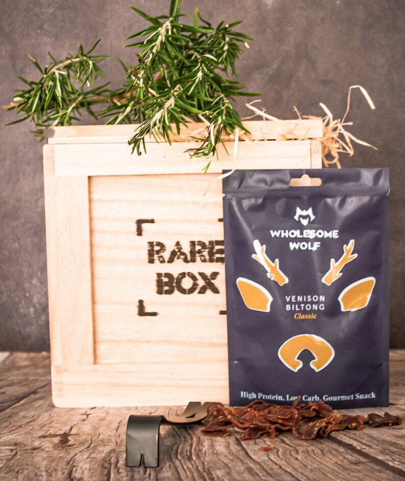 May include: A wooden crate with the words "Rare Box" printed on the front. The crate contains a bag of venison biltong from the brand "Wholesome Wolf". The bag is blue with a deer head logo and the text "Venison Biltong Classic". The biltong is also visible outside of the bag.