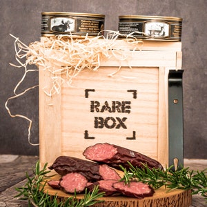 May include: A wooden box with the words "RARE BOX" printed on the front. The box is filled with shredded paper and contains two cans of canned elk and moose meat. There is also a wooden cutting board with sliced smoked sausage and rosemary sprigs.