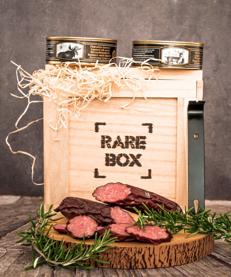 May include: A wooden box with the words "RARE BOX" printed on the front. The box is filled with shredded paper and contains two cans of canned elk and moose meat. There is also a wooden cutting board with sliced smoked sausage and rosemary sprigs.
